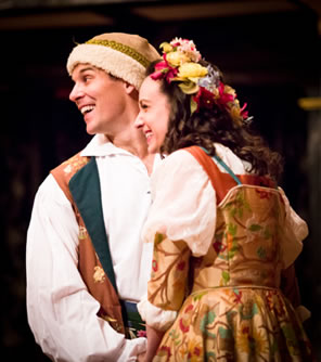 Florizel in white shirt, tan, fur-lined stocking cap, and brown and blue floral dress holds hands with Perdita in a floral tapestry dress, bulging-sleeved white blouse, and flowers in her hair